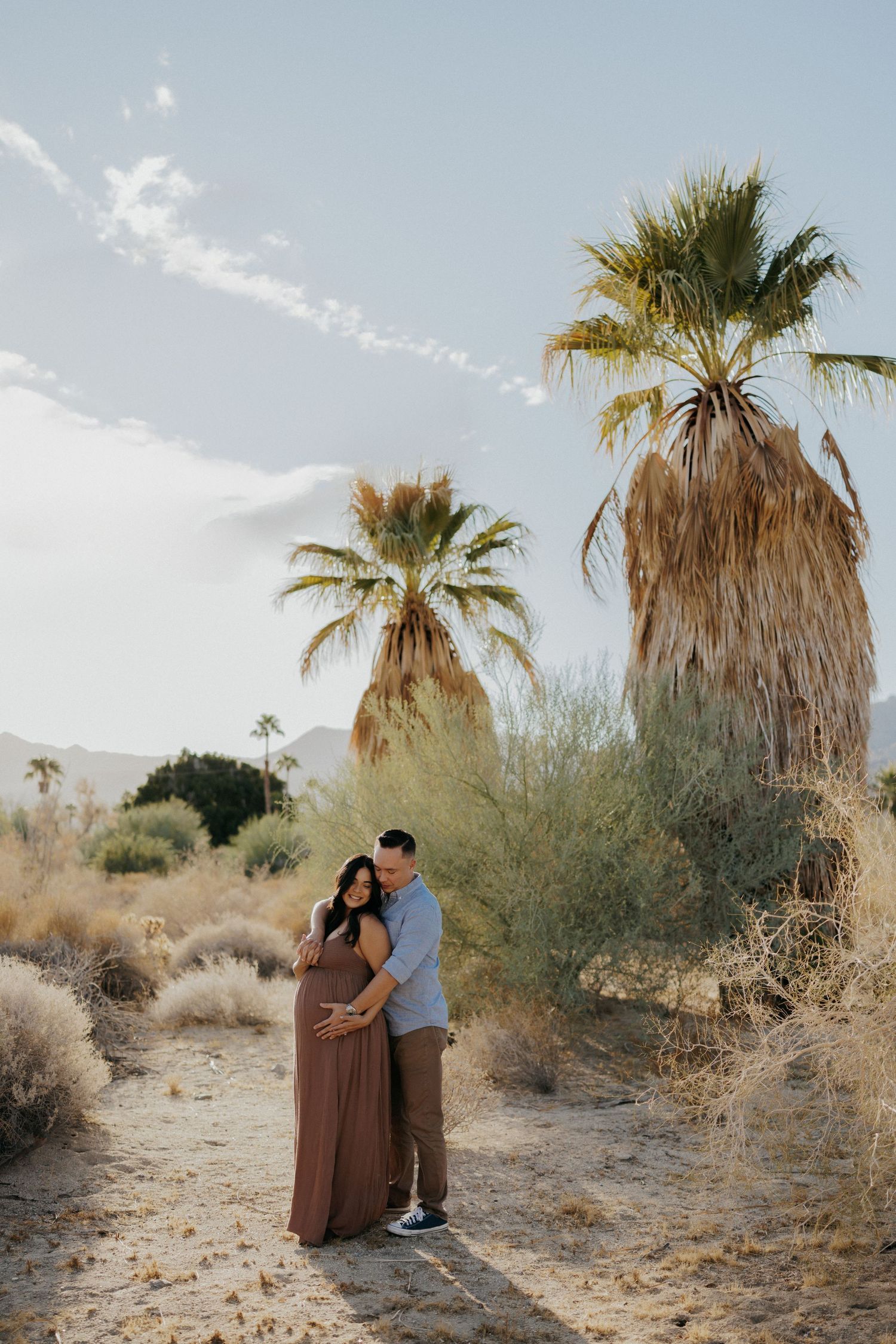 Couple poses in desert landscape with palm trees during maternity photo session in warm sunlight.