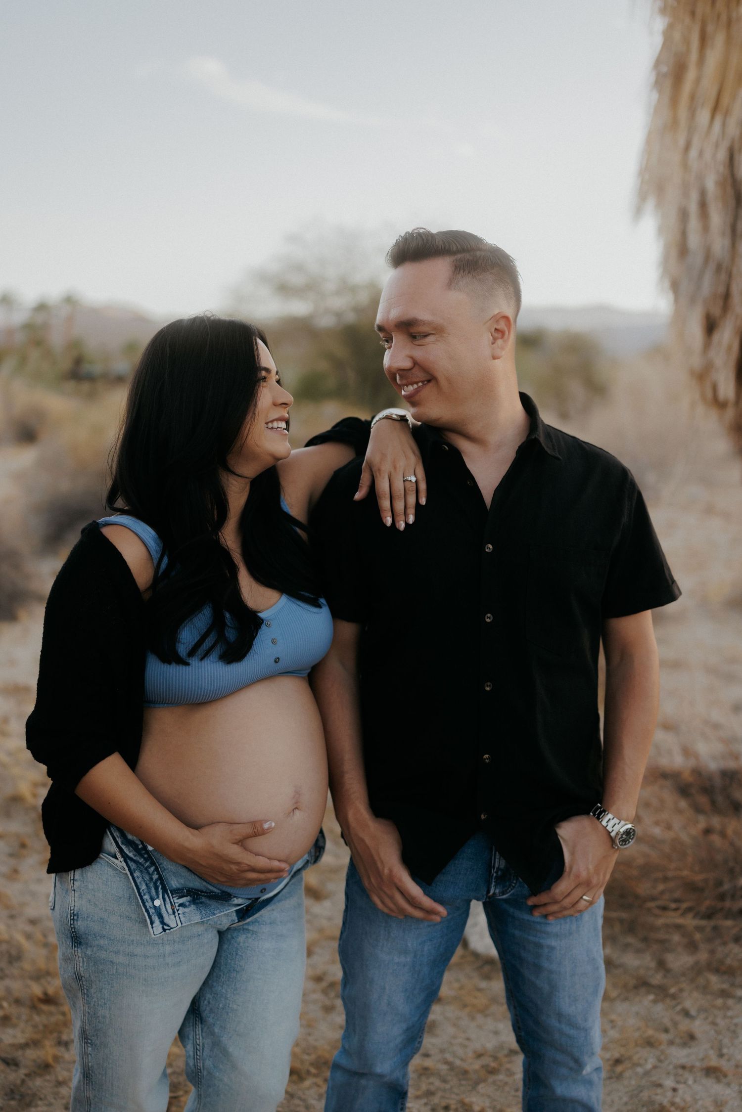 Smiling pregnant couple in casual black and blue outfits during outdoor maternity shoot in desert setting.