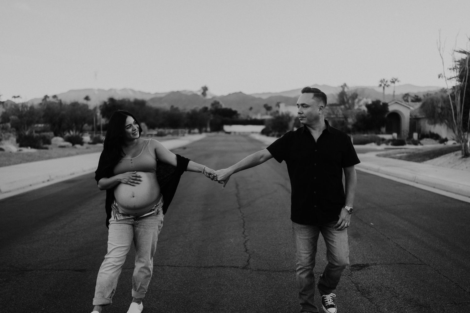 Black and white maternity photo of pregnant couple holding hands on empty road with mountains behind.