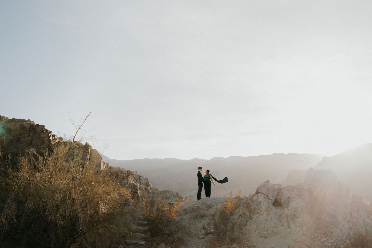Couple standing on rocky mountain ridge at golden hour with misty mountain range backdrop.