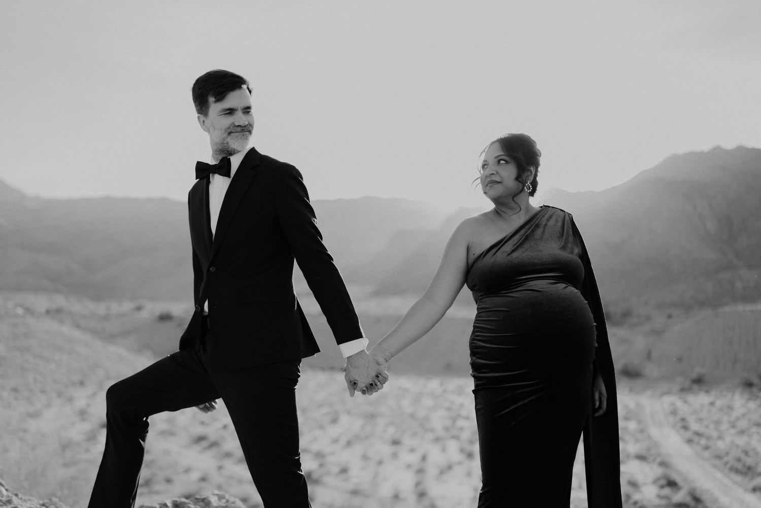 Black and white photo of a smiling pregnant couple holding hands in formal wear with desert mountains backdrop.