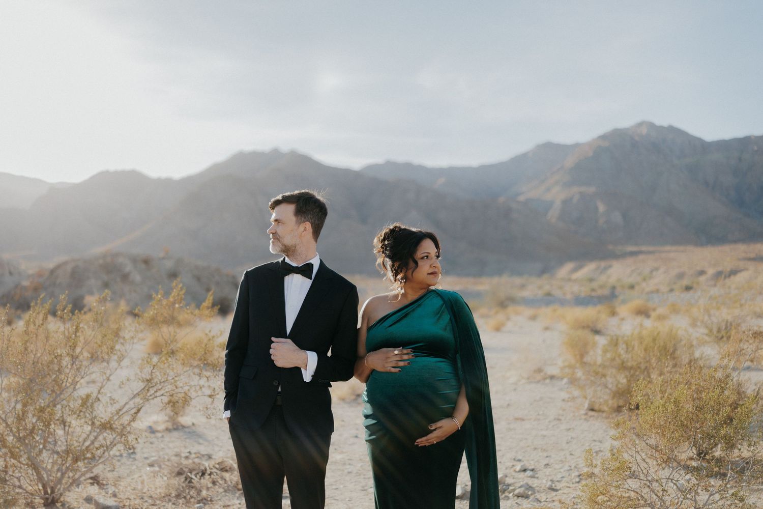 Pregnant couple in desert mountains, man in tuxedo, woman in emerald green gown, maternity photo session.