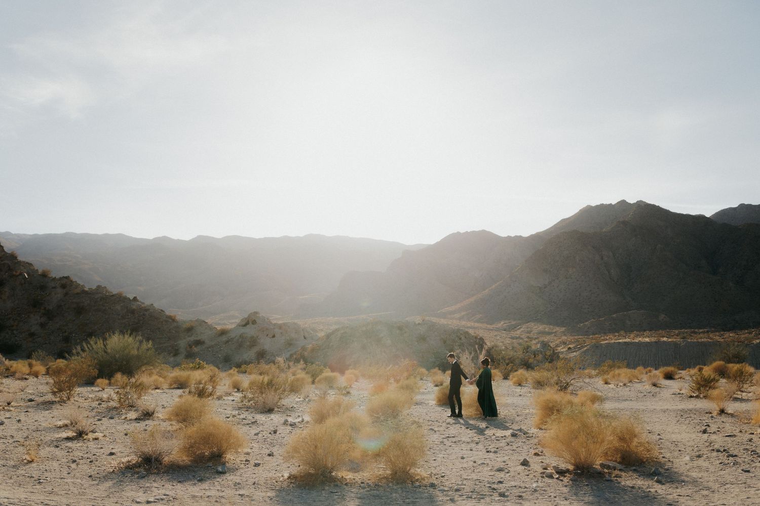 Two figures stand in a vast desert landscape with mountains and golden sunlight in the background.