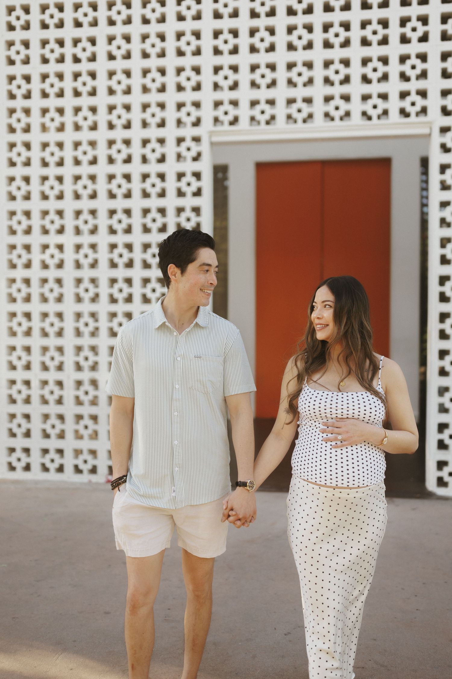 A smiling couple holds hands in front of a decorative white lattice wall, with the woman in a polka dot dress cradling her baby bump.