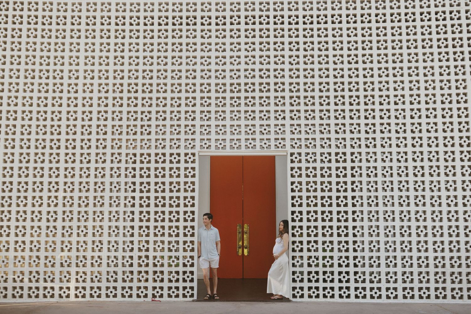 Two people in white outfits stand beside a red door set in a decorative geometric patterned white wall.