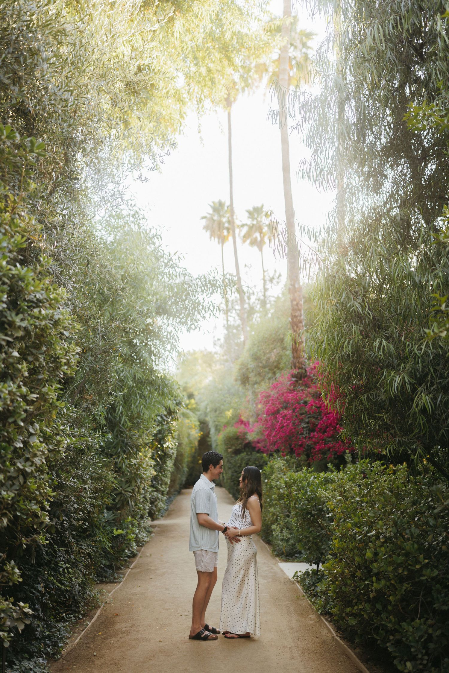 Couple holding hands on a sunlit garden path surrounded by lush greenery and pink bougainvillea blooms.
