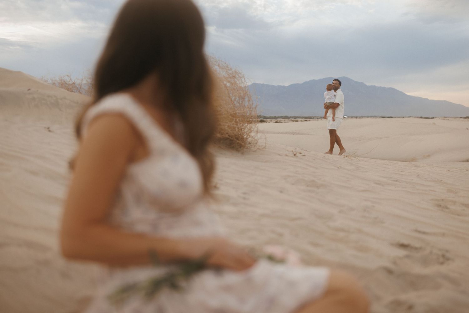 Couple in white outfits on sandy desert dunes with mountain backdrop, romantic engagement photo session.