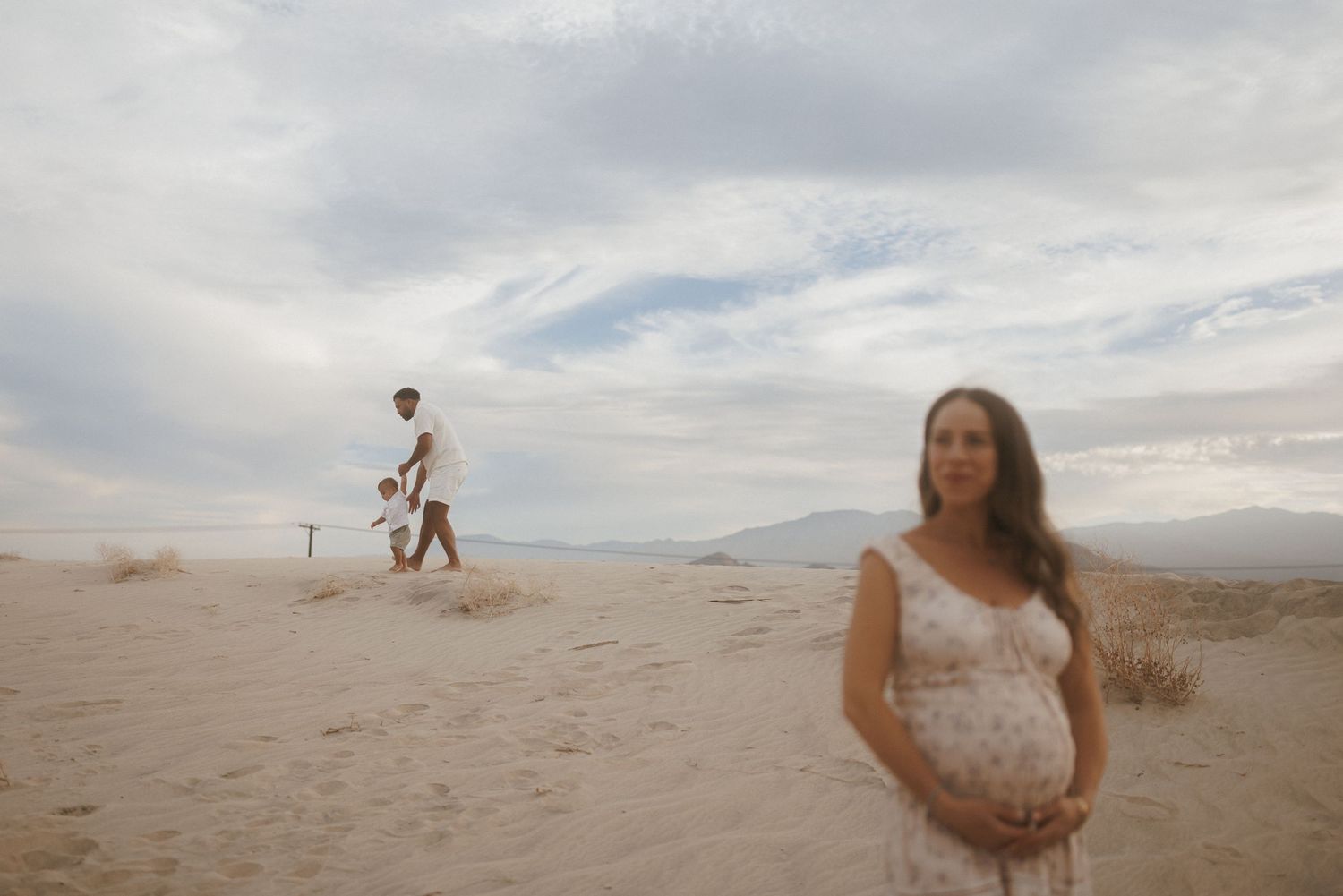Pregnant woman in white dress poses on sandy desert dunes with family in background under cloudy sky.