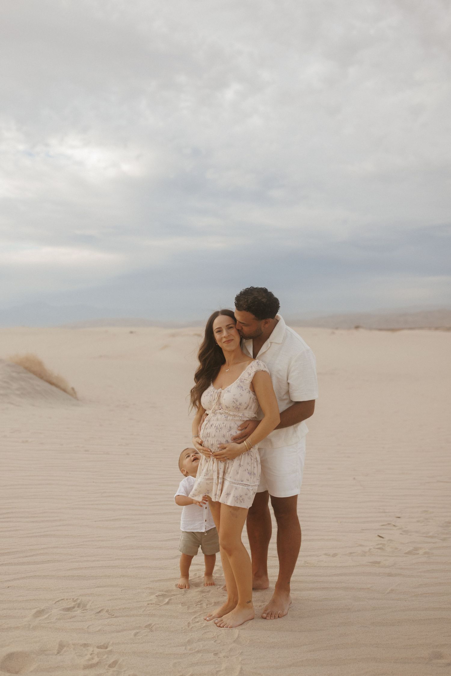 Pregnant couple with toddler posing on sandy desert dunes during a maternity photoshoot under cloudy skies.