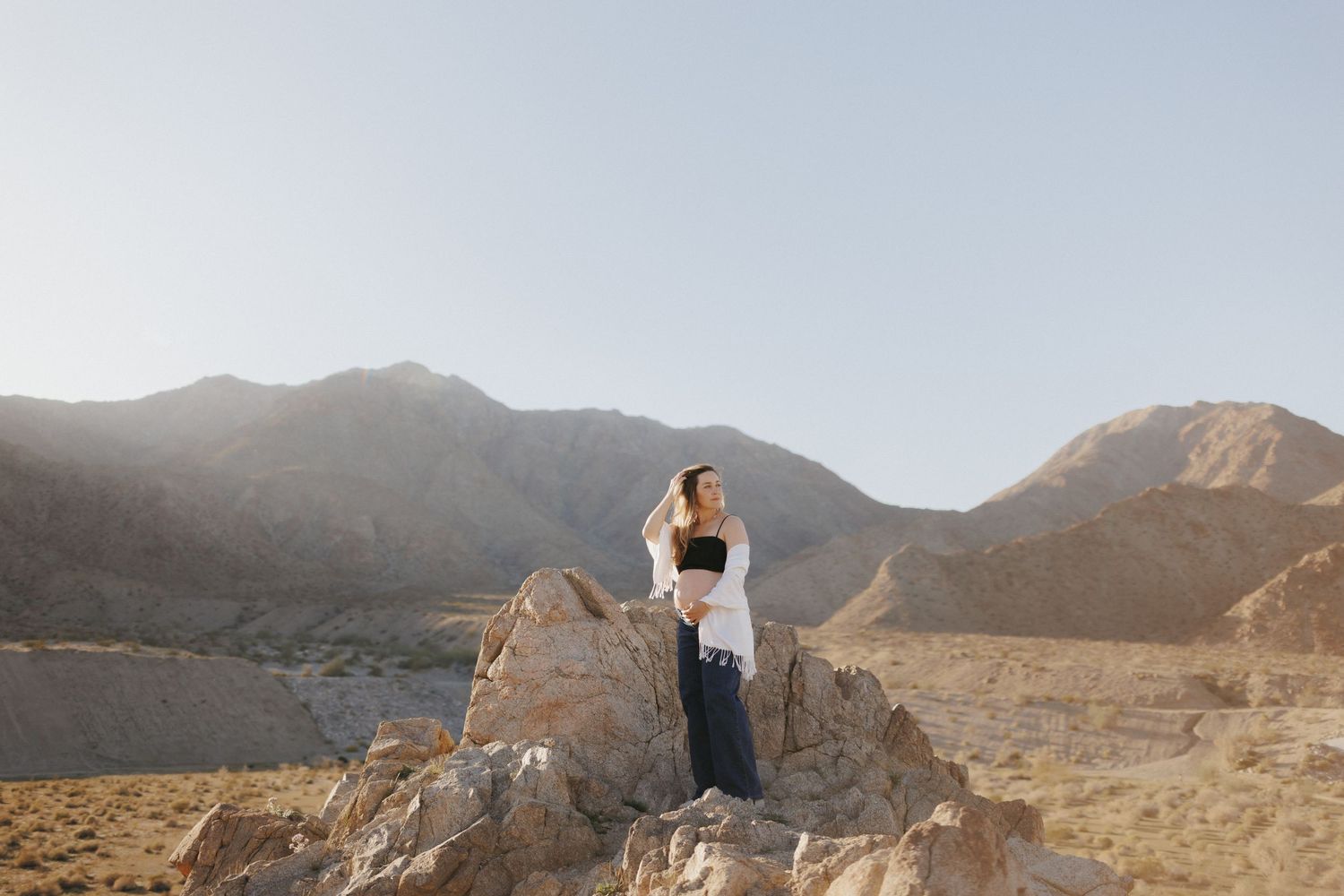 Woman standing on rocks in a desert landscape with mountains and golden sunlight in the background.