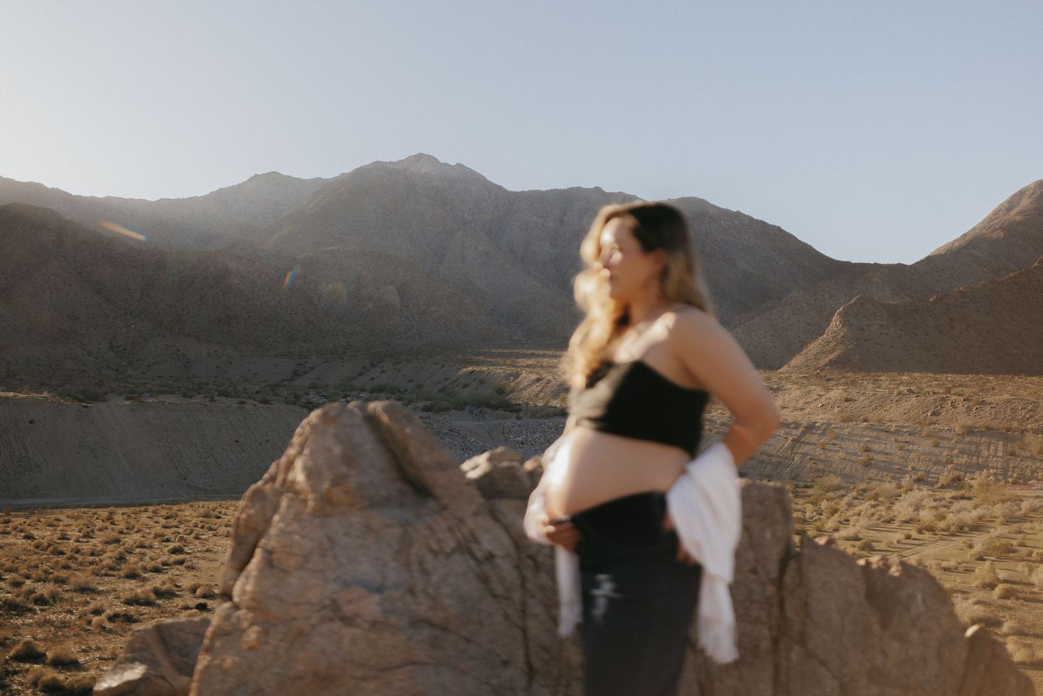 Pregnant woman in black dress standing on desert rocks with mountain backdrop in golden hour light.