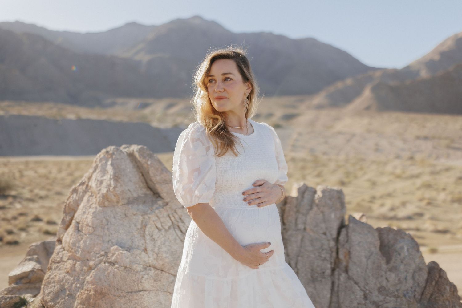Pregnant woman in white lace dress poses among desert rocks with mountains in background.
