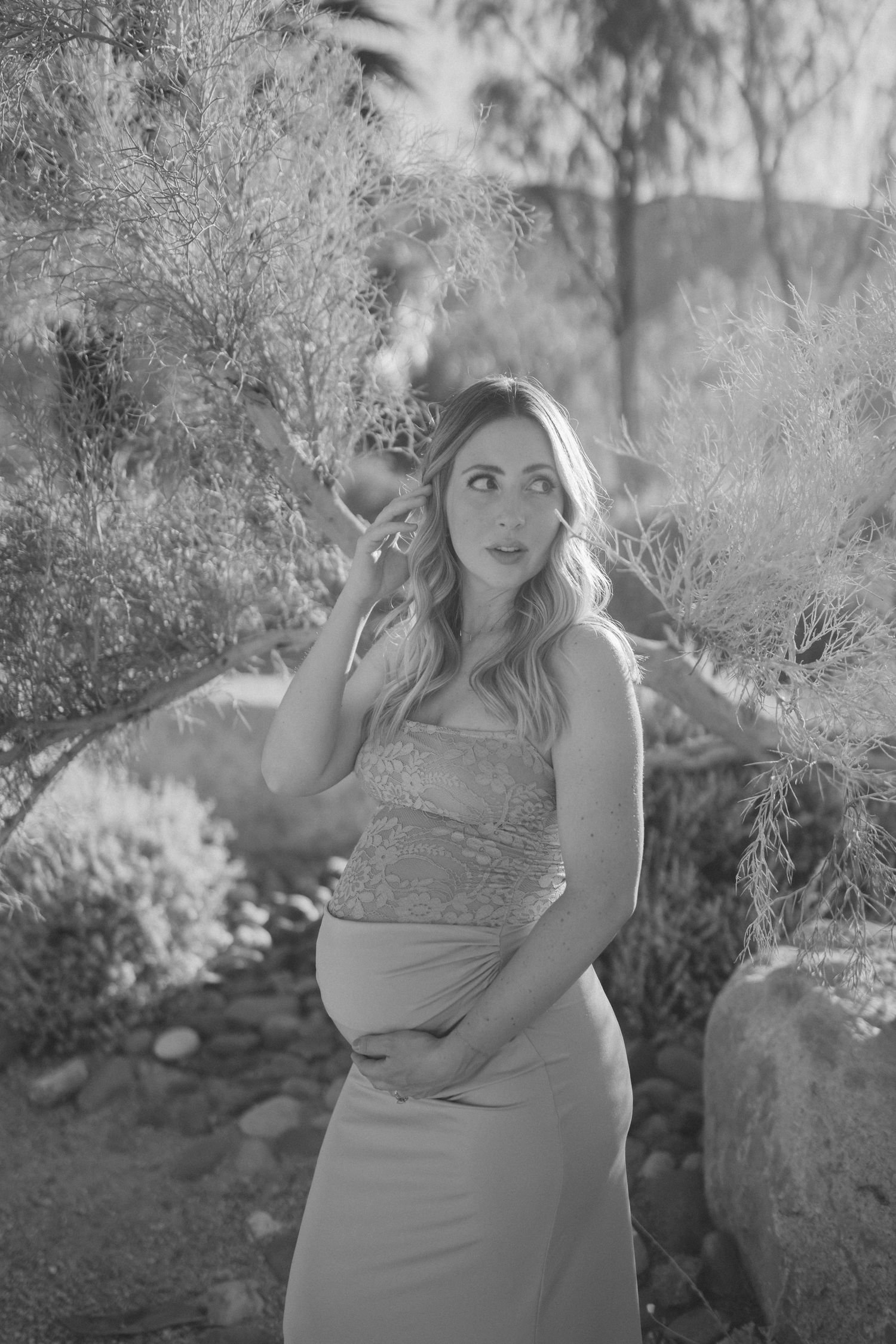 Black and white maternity photo of a pregnant woman in a sequin top and long skirt, posing outdoors among desert plants.