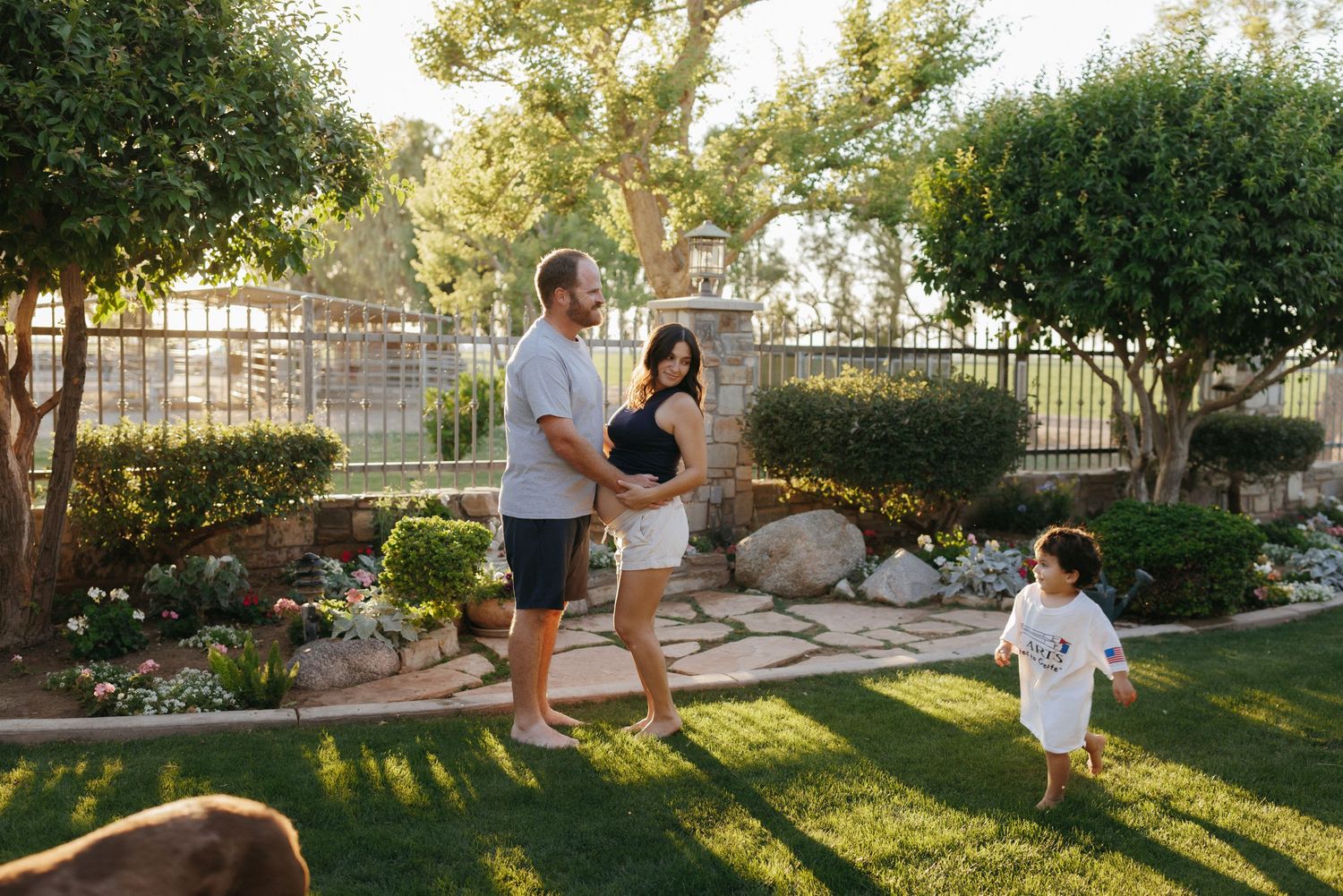 A family of three enjoys a sunny afternoon in a lush backyard garden, with a toddler running on green grass.