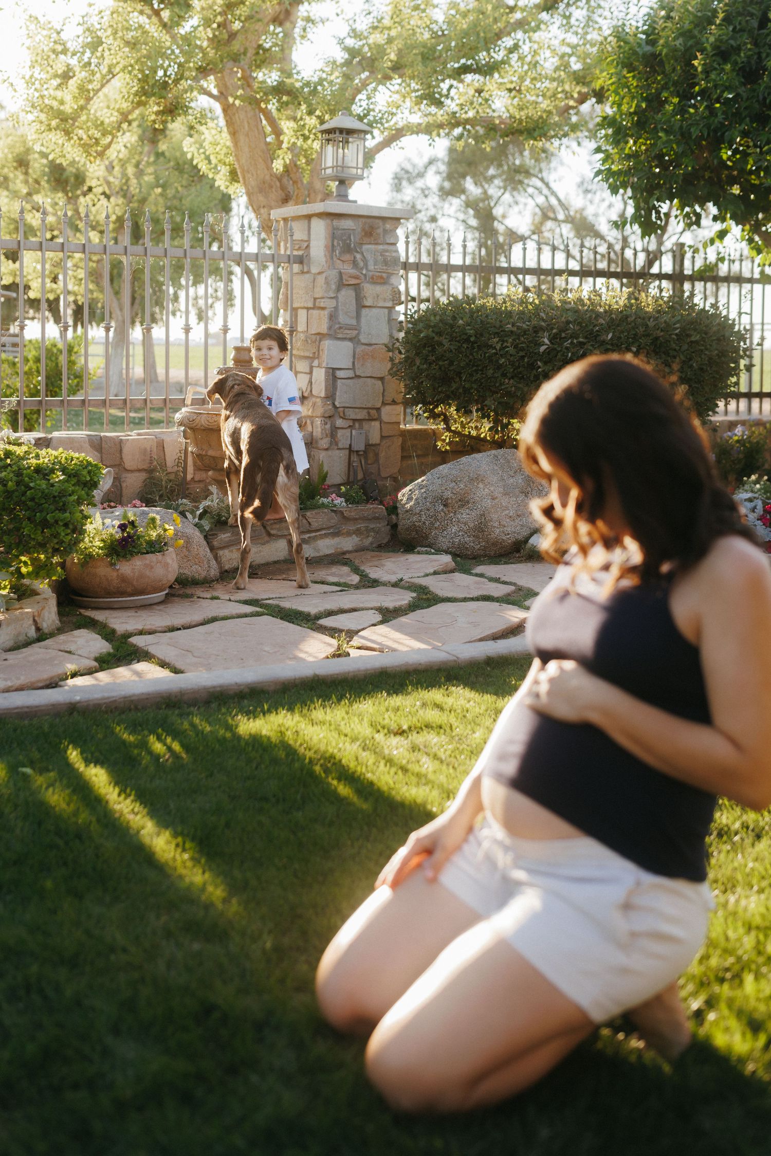 Pregnant woman kneeling on grass in backyard, child playing with dog near stone gate in golden sunlight.