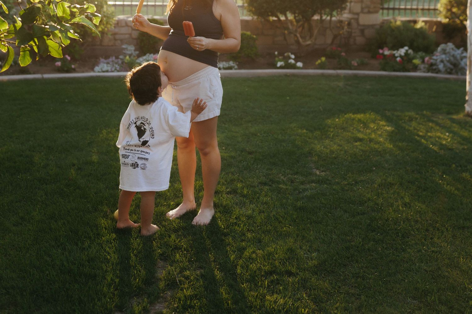 A toddler in a white shirt stands beside an adult on a green lawn during golden hour sunset light.