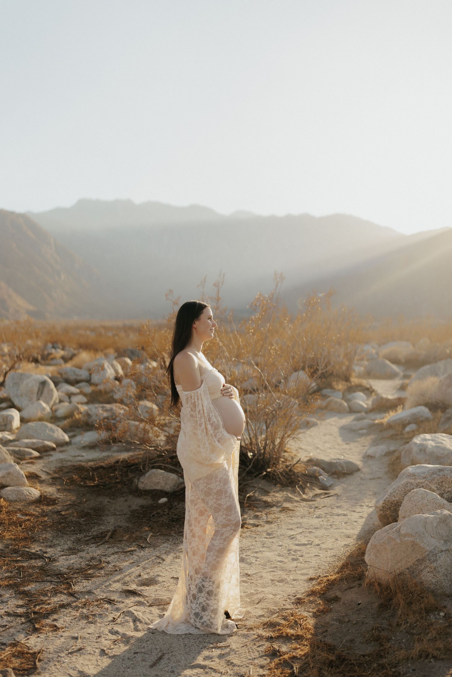 Pregnant woman in white lace gown cradling baby bump in desert landscape with mountains and golden sunlight.