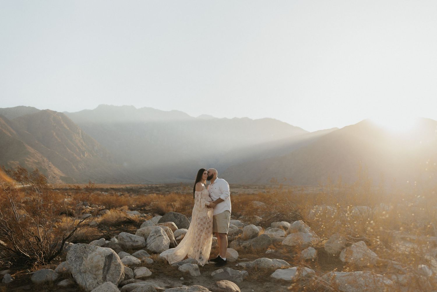 Couple embracing on rocky desert landscape with golden sunset and mountain backdrop.