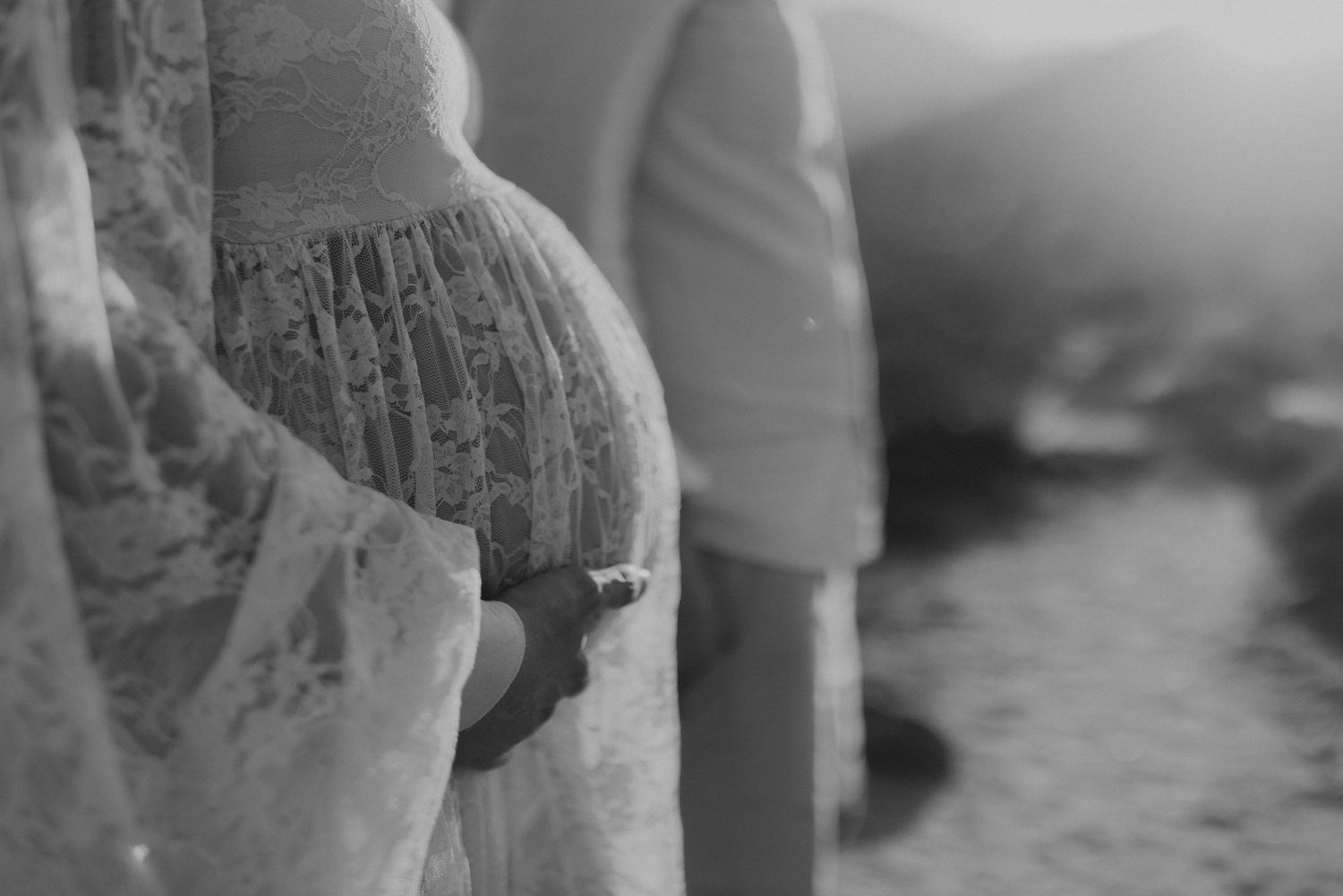 Black and white close-up of a pregnant woman in lace dress holding her baby bump outdoors.