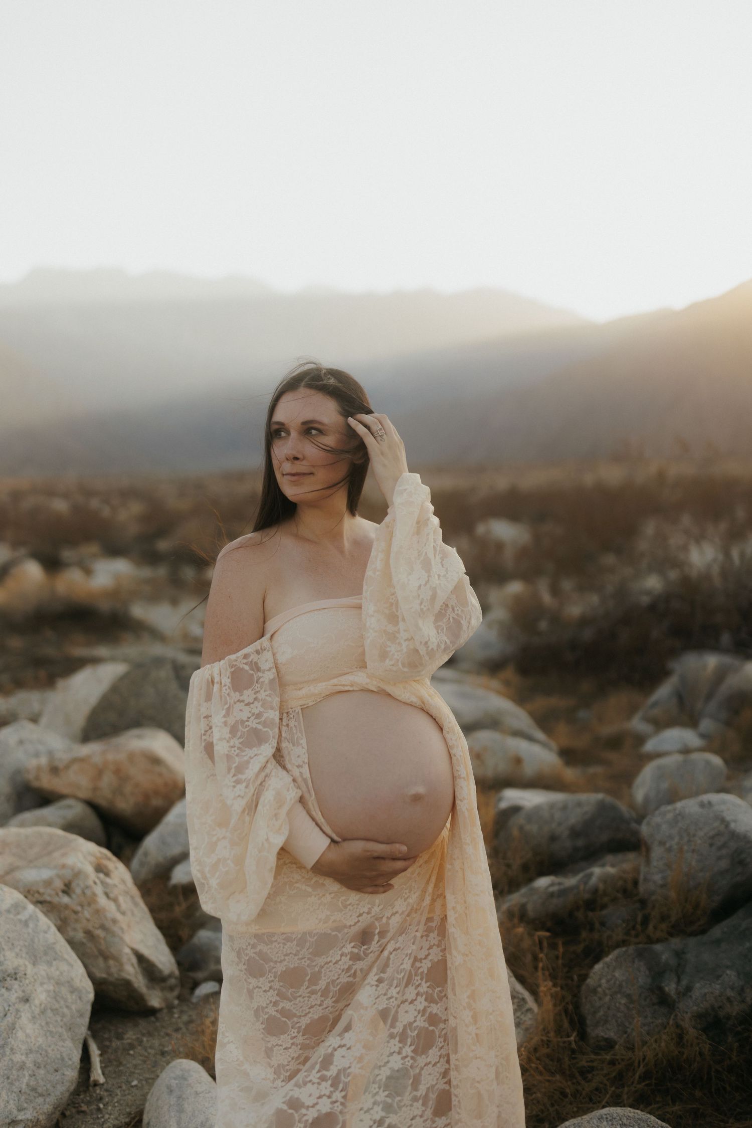 Pregnant woman in lace robe cradling baby bump at golden hour with rocky desert landscape and mountains.