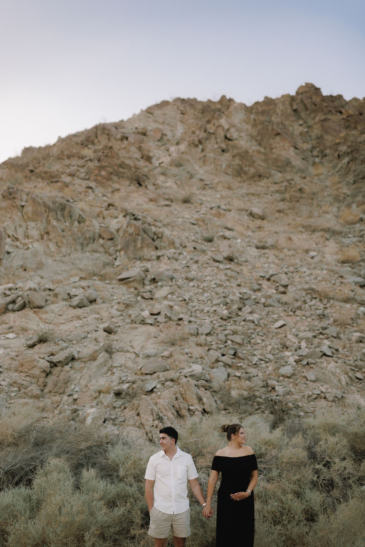 Couple holding hands in desert landscape with rocky mountain backdrop, woman in black dress, man in white shirt.