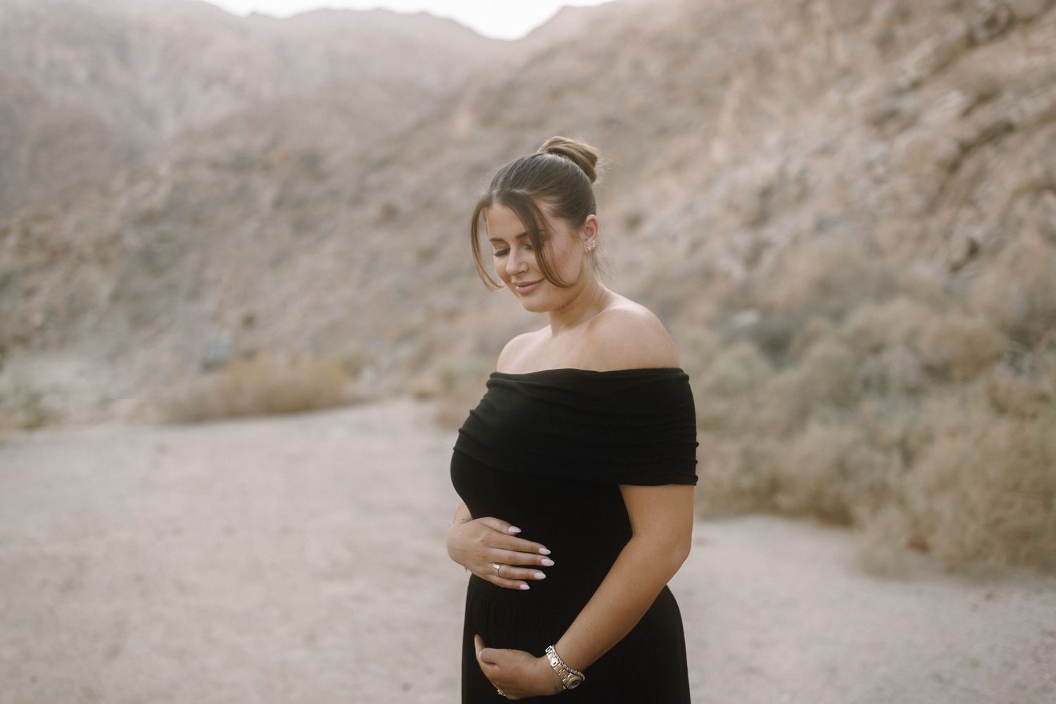 Pregnant woman in black off-shoulder dress cradling her baby bump outdoors in a desert canyon setting.