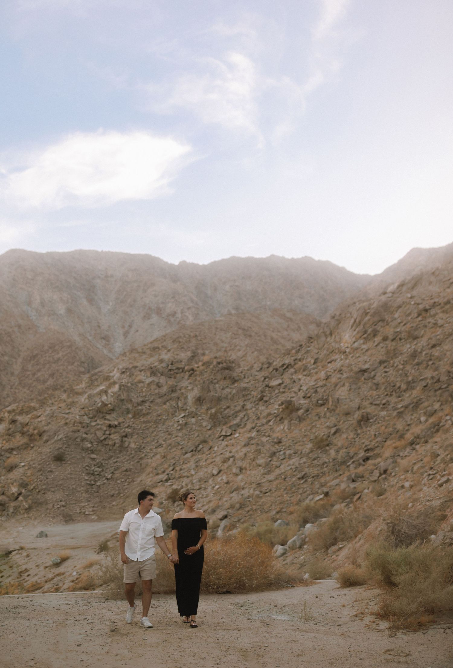 Couple holding hands walking on a desert trail with dramatic rocky mountains in the background under a hazy sky.