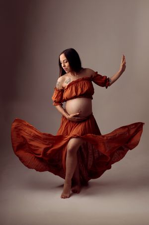 Pregnant woman in flowing rust orange off-shoulder dress twirling gracefully in studio maternity shoot.