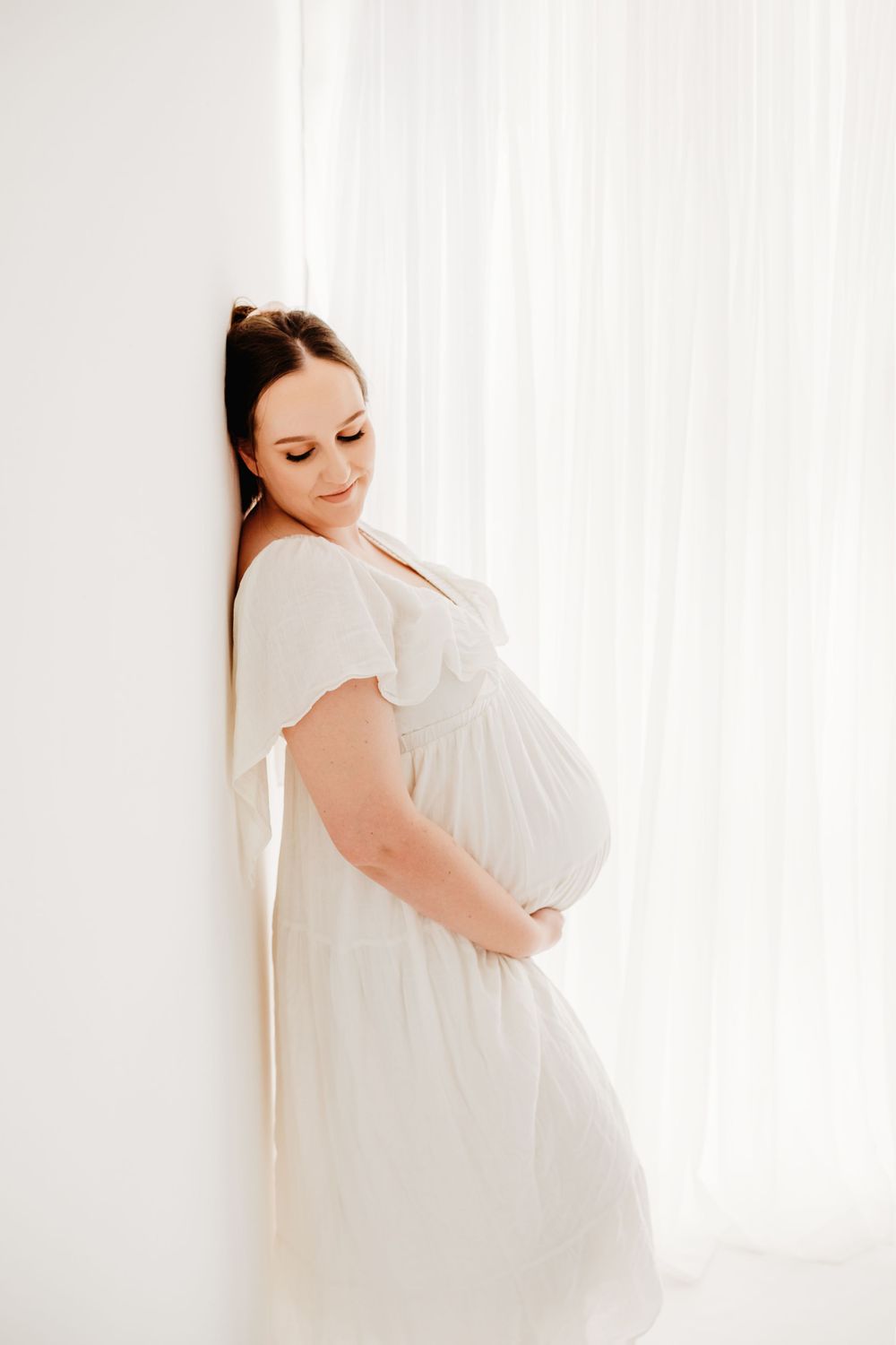 Expectant mother in white dress poses against white curtain backdrop in soft maternity photoshoot.