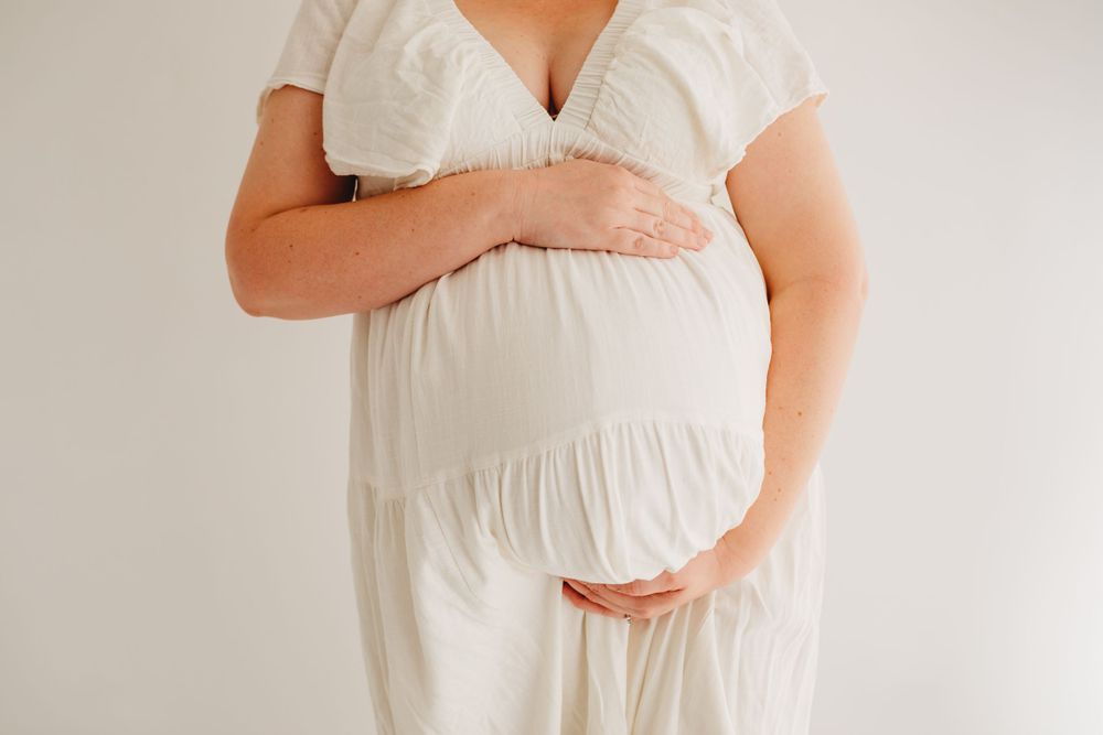 Pregnant woman in white dress cradles baby bump during indoor maternity session.
