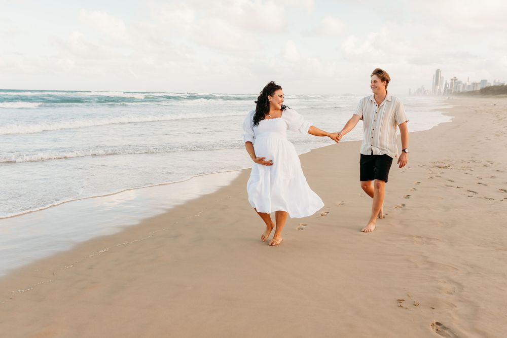 Couple in white outfits playfully running along beach shoreline at sunset.