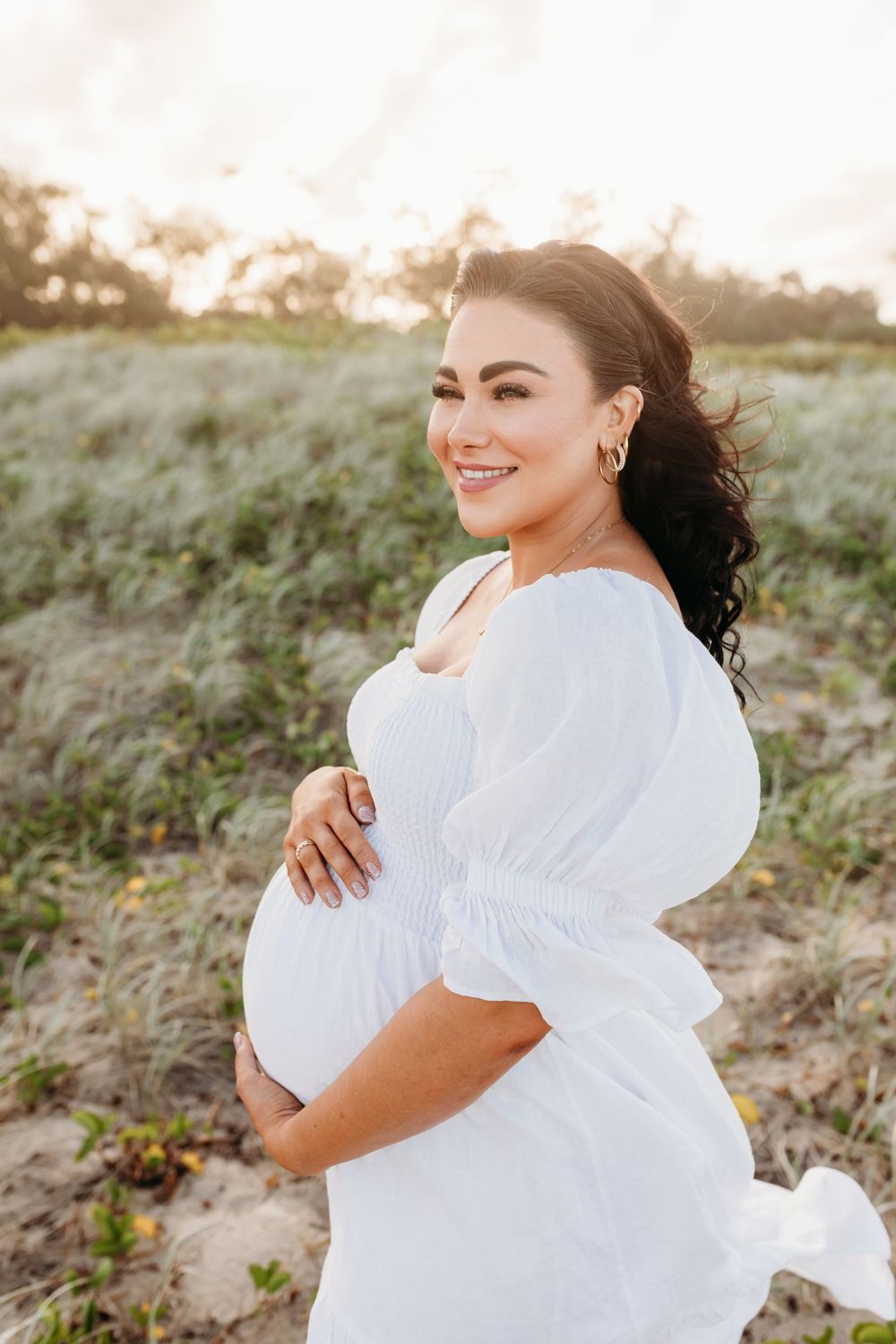 Maternity photoshoot at beach featuring woman in flowing white dress at sunset.