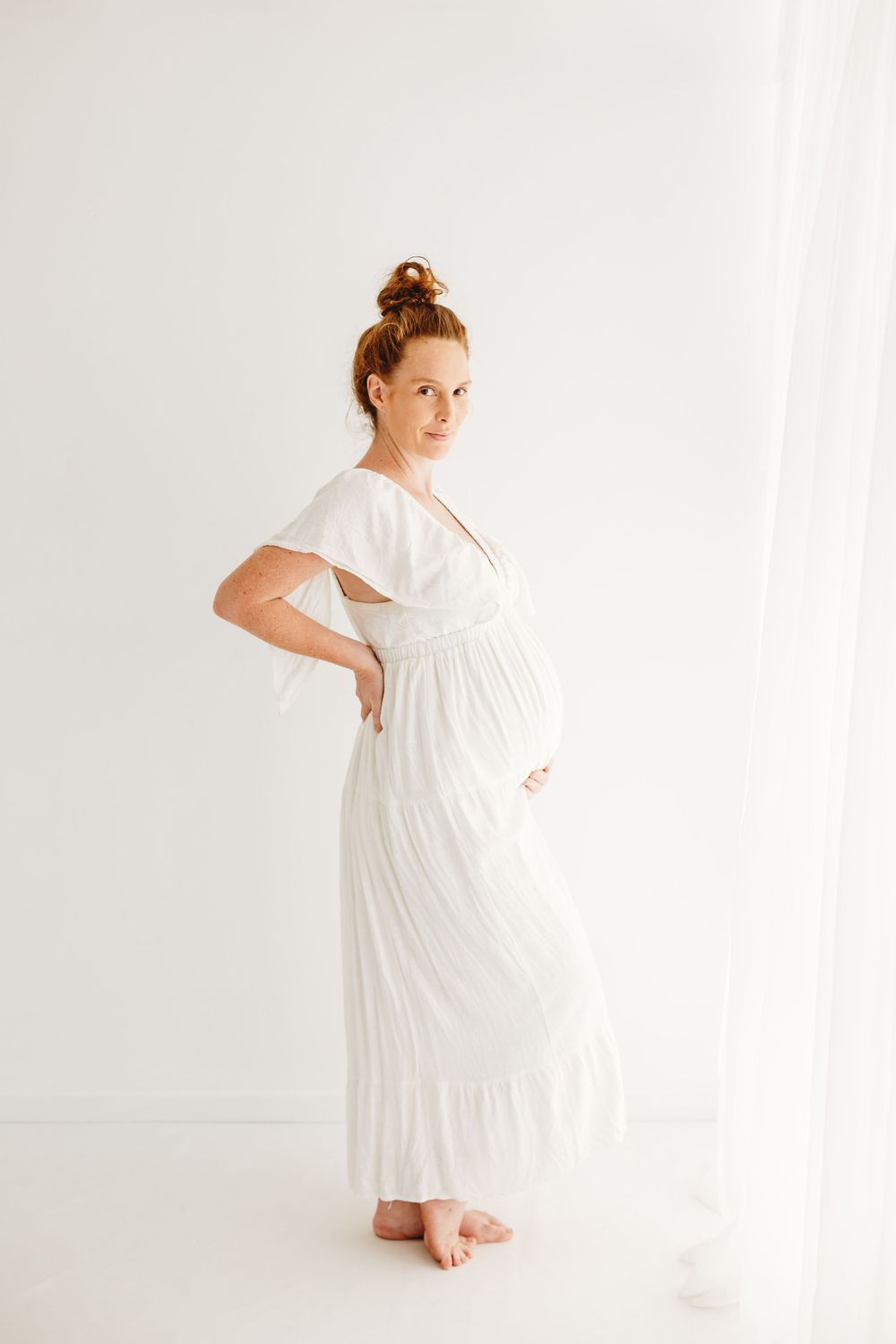 Pregnant woman in white dress stands sideways showing her baby bump against light background.