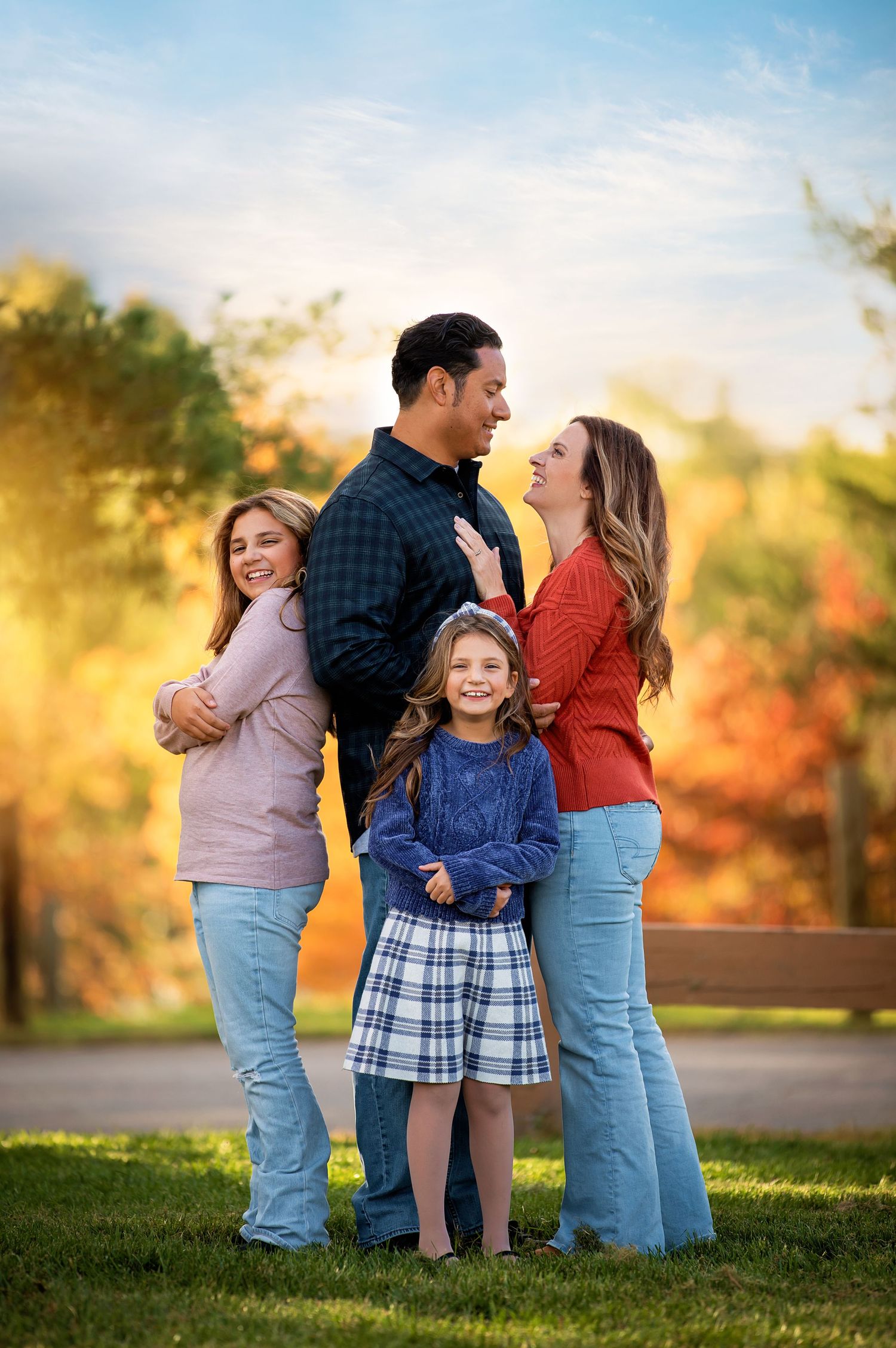 A family of four poses together outdoors during autumn with colorful fall foliage in the background.