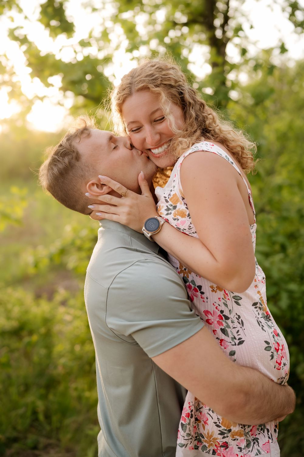 A man lifts and kisses a smiling woman in a floral dress outdoors, bathed in warm golden sunset light.