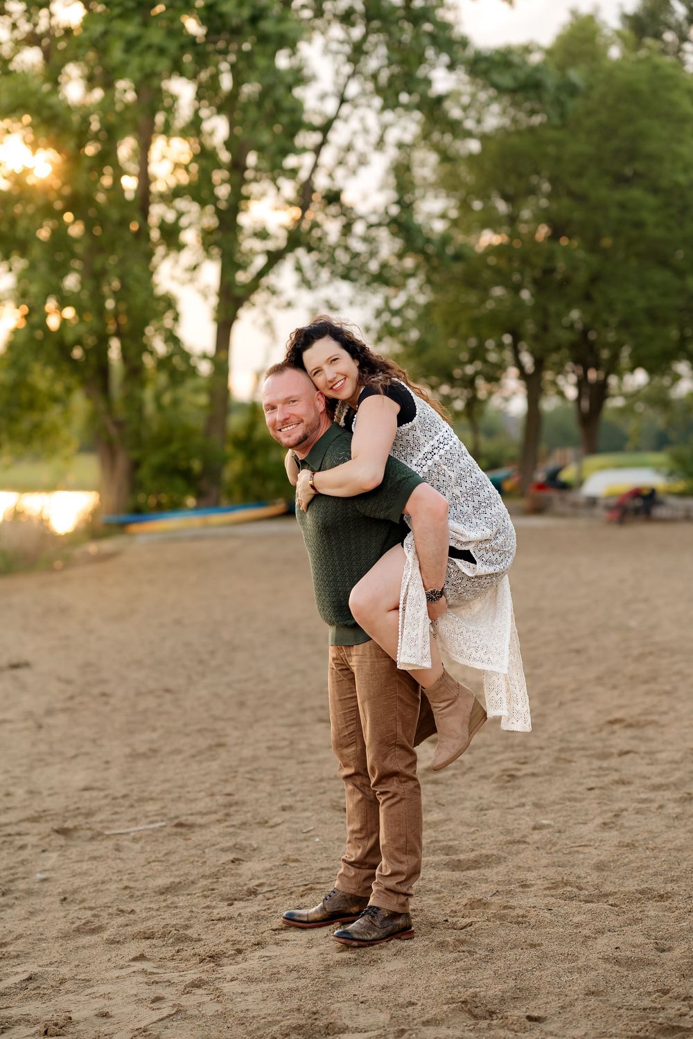 A happy couple enjoys a piggyback ride on a sandy beach at sunset with lush green trees in the background.
