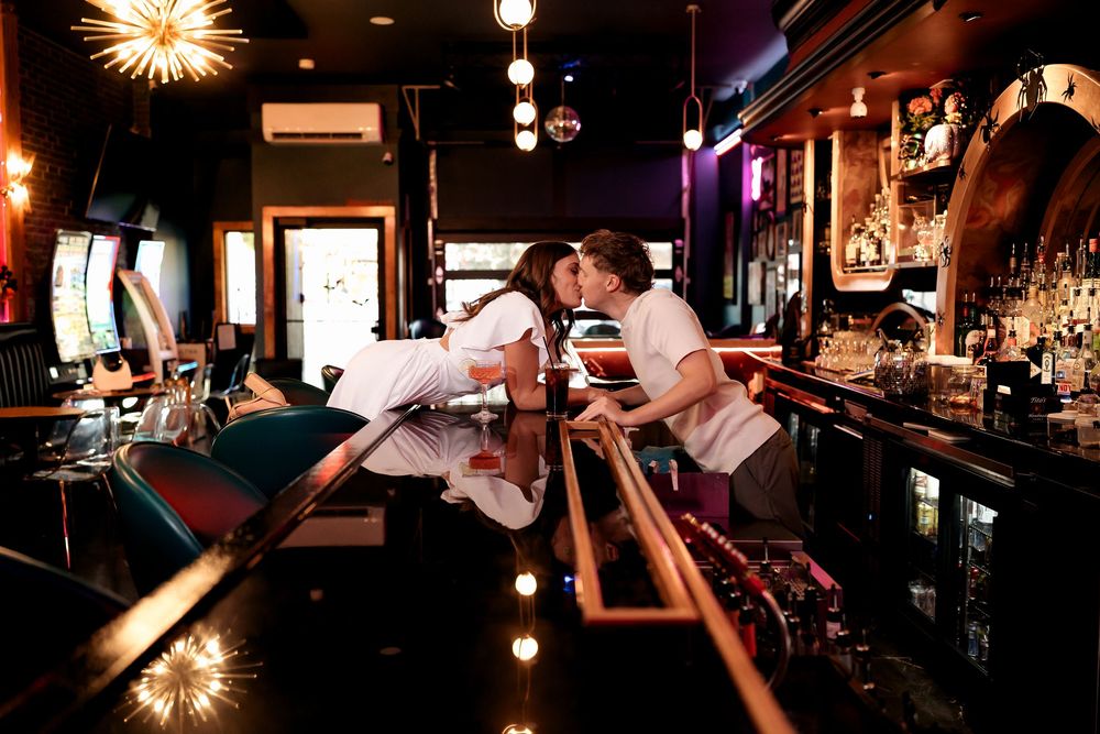 Two bartenders in white uniforms lean over a polished bar counter in a dimly lit establishment with warm ambient lighting.