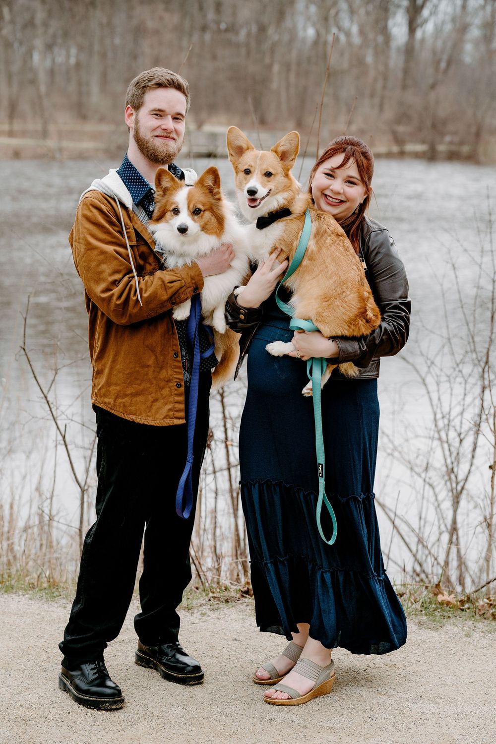 A smiling couple holds two Corgi dogs outdoors near a pond, the woman in a navy dress and man in a brown jacket.