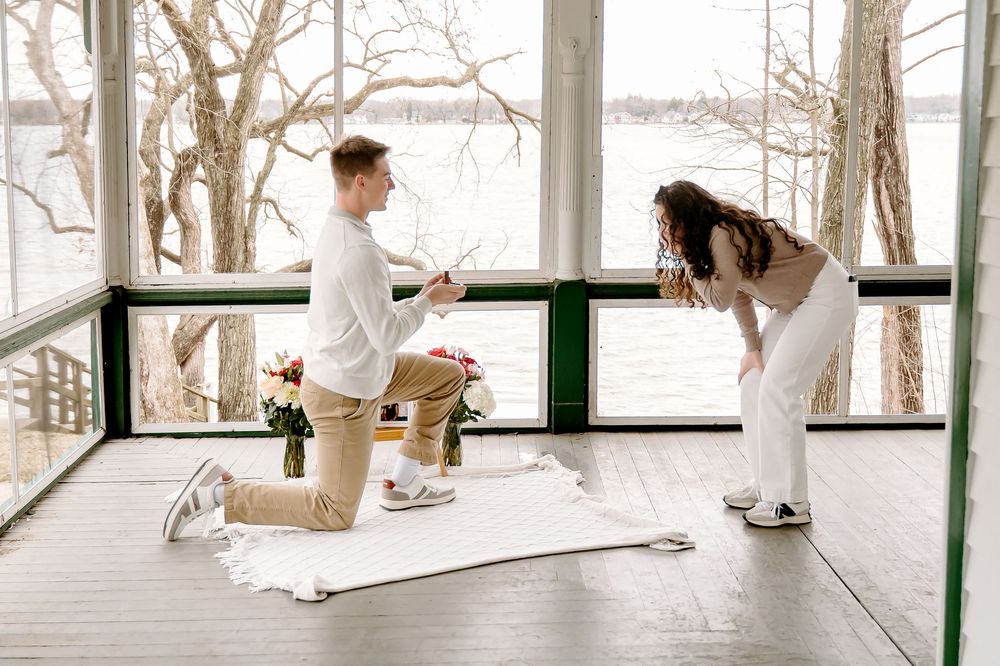Man on one knee proposing to a woman on a sunlit porch overlooking a lake, with flowers nearby.