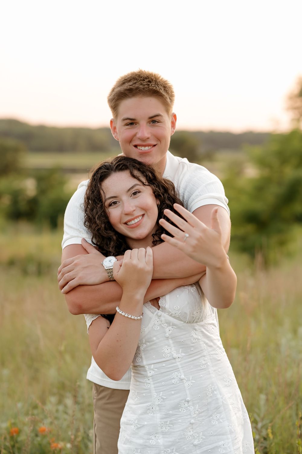 A young couple embraces in a sunlit field, the woman in white showing an engagement ring while smiling joyfully.