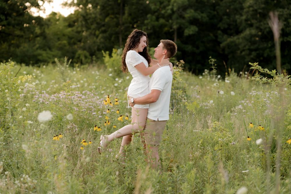Couple embracing in a wildflower meadow during golden hour, pregnant woman lifted by partner outdoors.