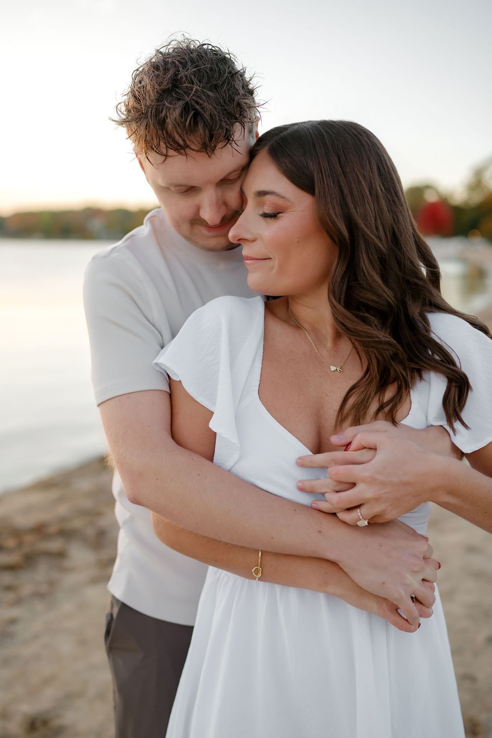 A romantic couple embraces on a beach at sunset, with the man holding the woman from behind in a tender moment.