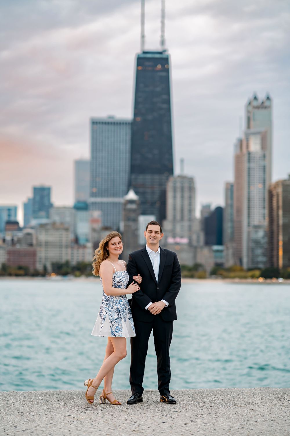 Couple poses for engagement photos along Lake Michigan with Chicago's iconic skyline and John Hancock building in background.