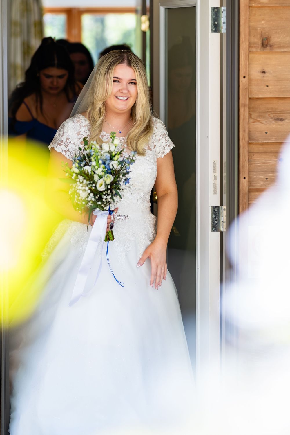 Someone in a delicate lace wedding dress holds a beautiful blue and white bouquet while standing in a doorway.