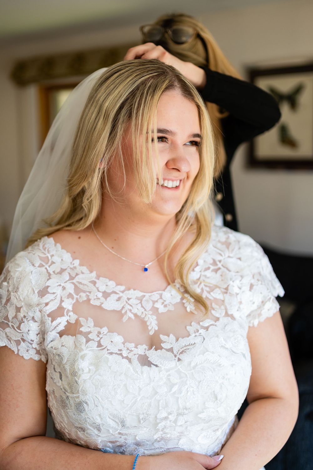 A bride wearing a white lace wedding dress with sheer details and a delicate neckline smiles during wedding preparations.