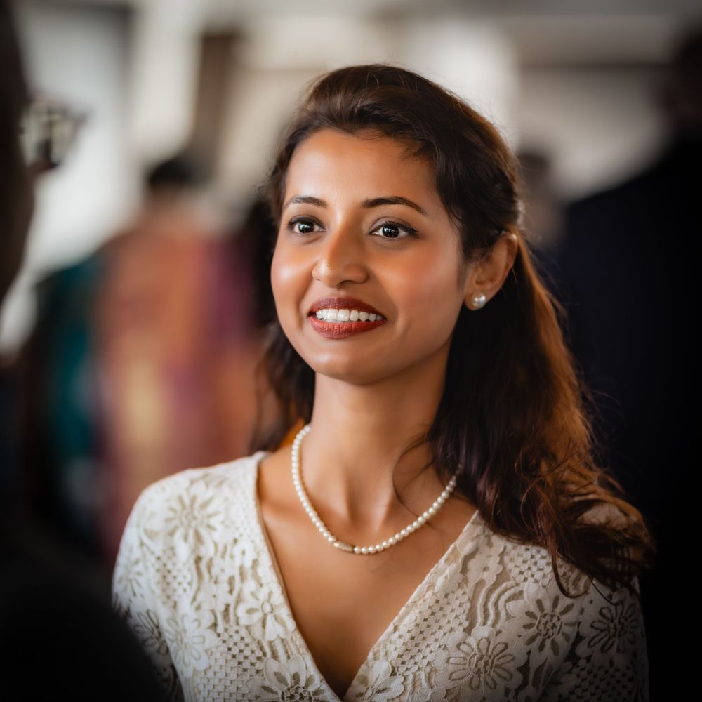Someone wearing a white lace dress and pearl necklace smiles warmly in an indoor setting.
