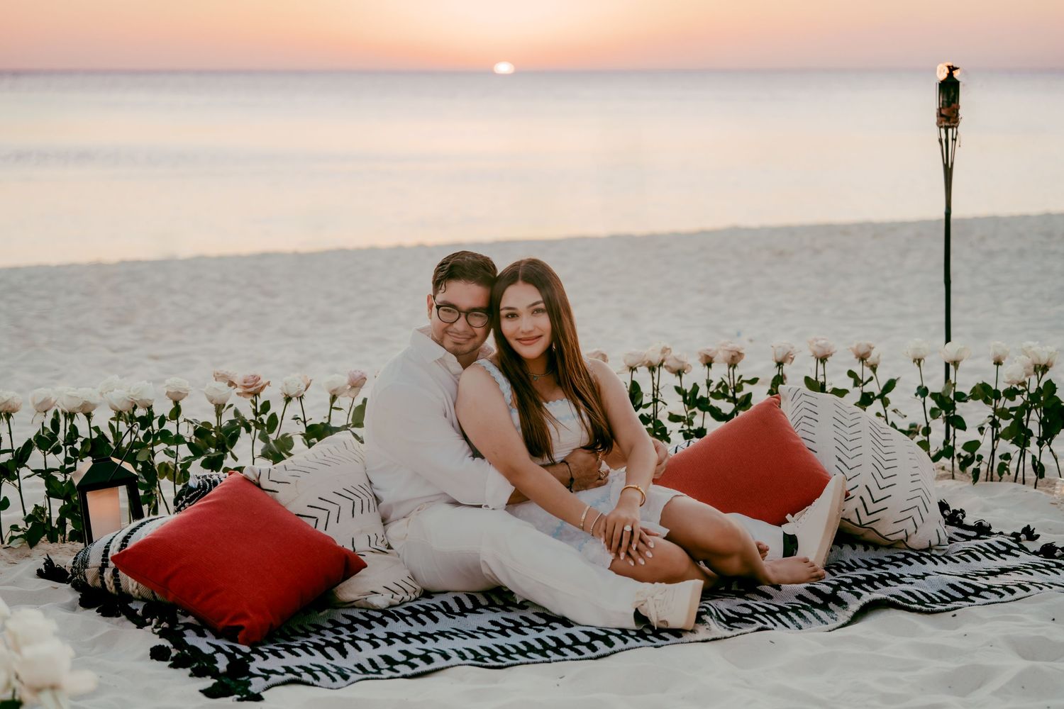Two people relax on a blanket with red pillows at sunset by the ocean with tropical plants nearby. Engagement photography Grand Cayman