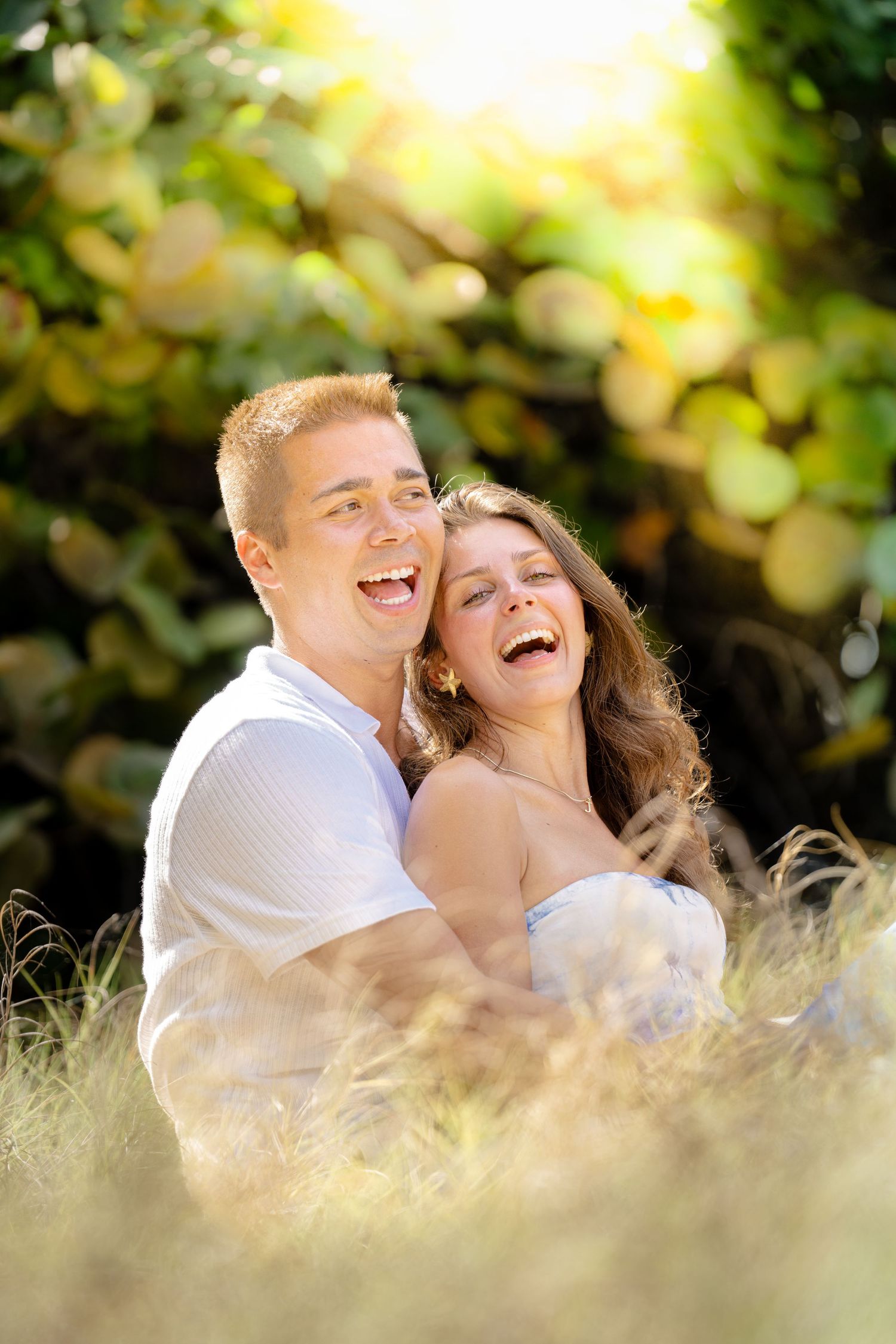 A joyful couple laughs together in a sunny meadow with soft bokeh and golden light during their outdoor portrait session.