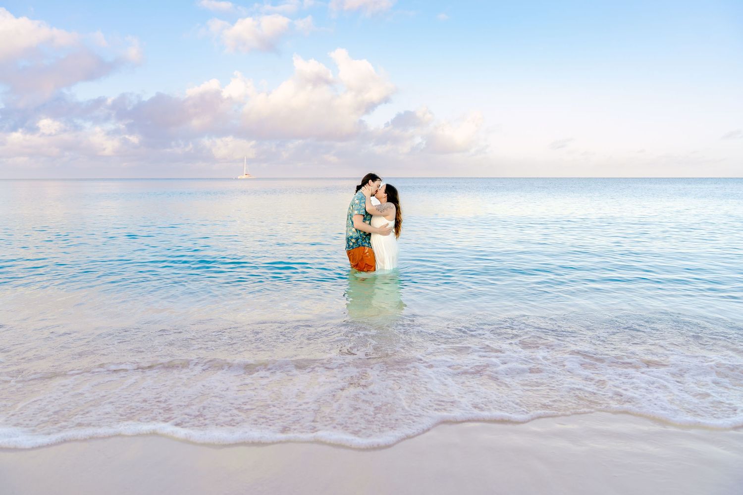 A couple embraces while standing in crystal clear turquoise ocean waters at sunrise along a tropical beach.