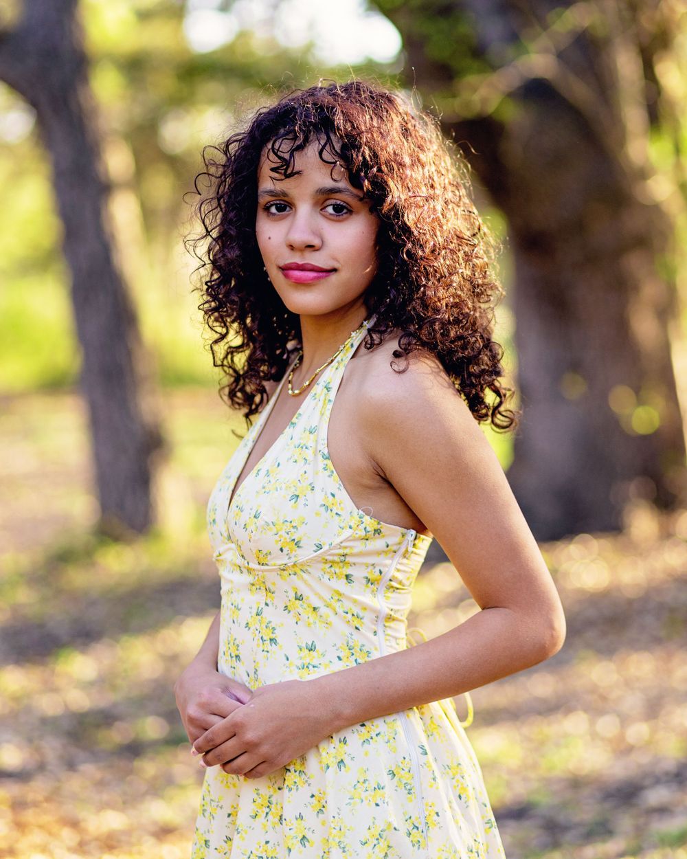 Person in yellow floral halter dress poses outdoors in nature with beautiful curly hair and natural lighting.